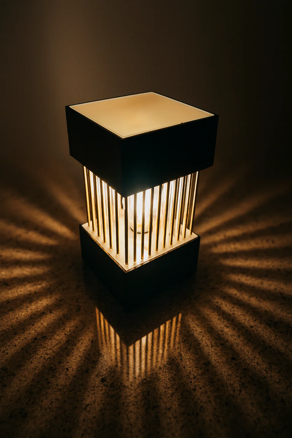 Warm leuchtende Lampe mit goldfarbenen senkrechten Stäben zwischen einem dunklen Sockel und einem quadratischen Oberteil